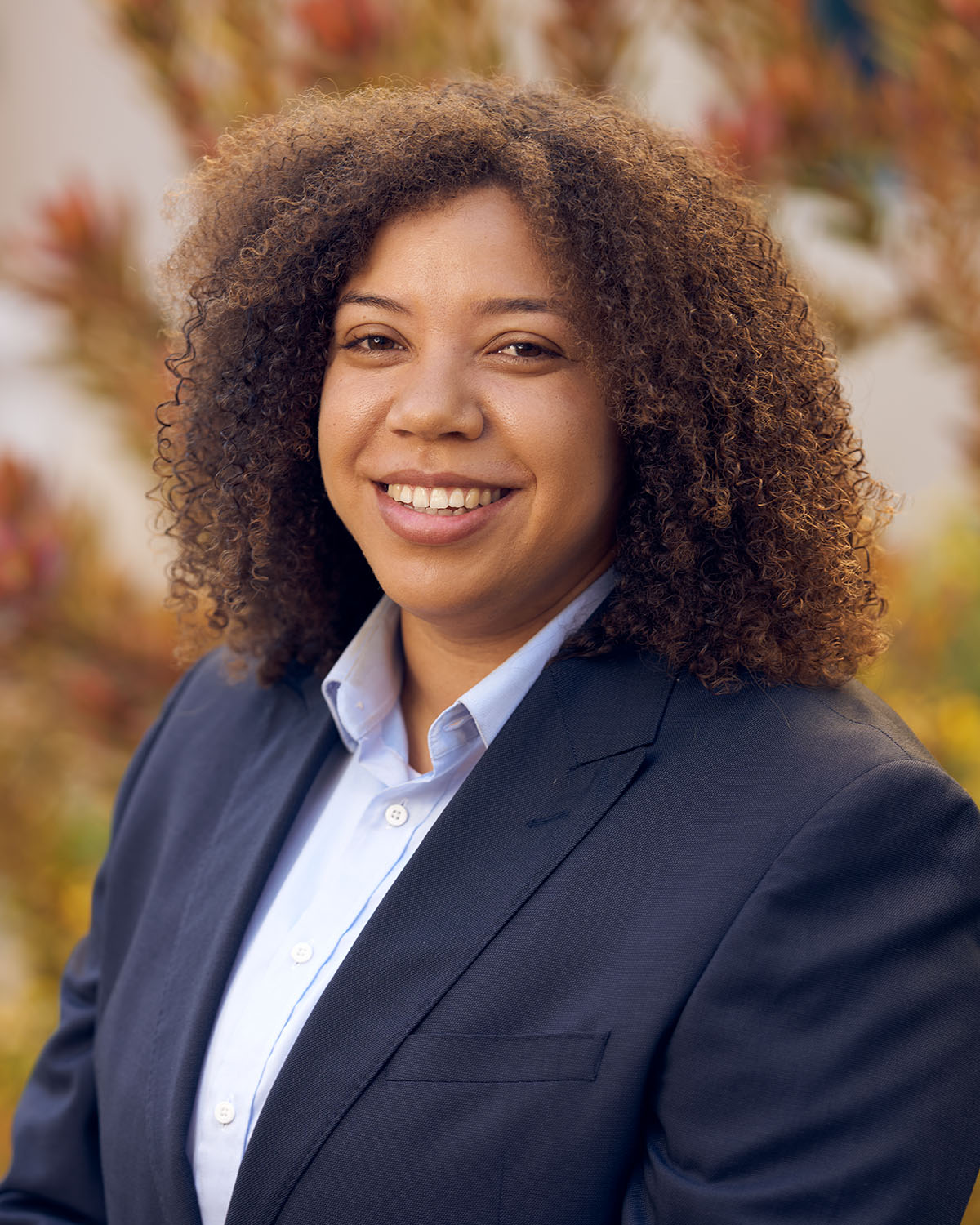 Corporate headshot of a woman with curly hair in a navy blazer smiling warmly outdoors in front of warm autumn foliage — Headshots by Guffy, San Diego