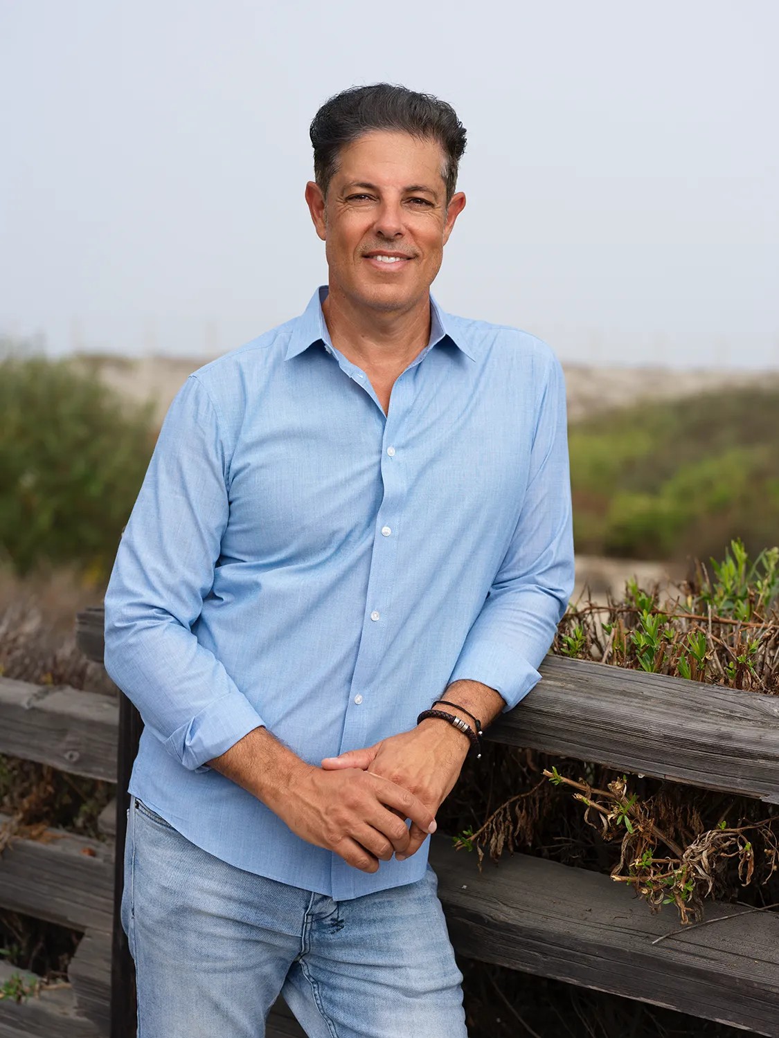 Personal branding headshot of a man in a light blue shirt leaning on a weathered wood railing at a coastal San Diego beach location — Headshots by Guffy, San Diego