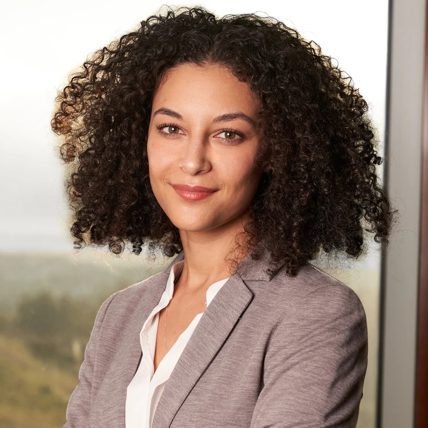 Professional headshot of a woman with natural curly hair in a gray blazer, photographed in front of a floor-to-ceiling window with a landscape view — Headshots by Guffy, San Diego