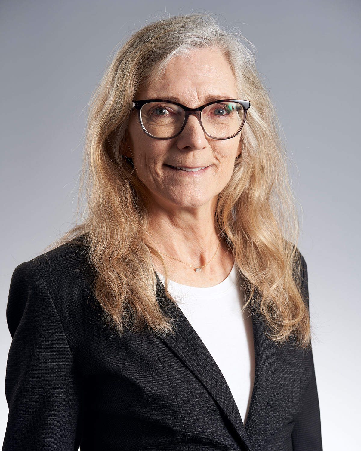 Professional headshot of a woman with silver hair and glasses in a black blazer against a gray gradient background — Headshots by Guffy, San Diego