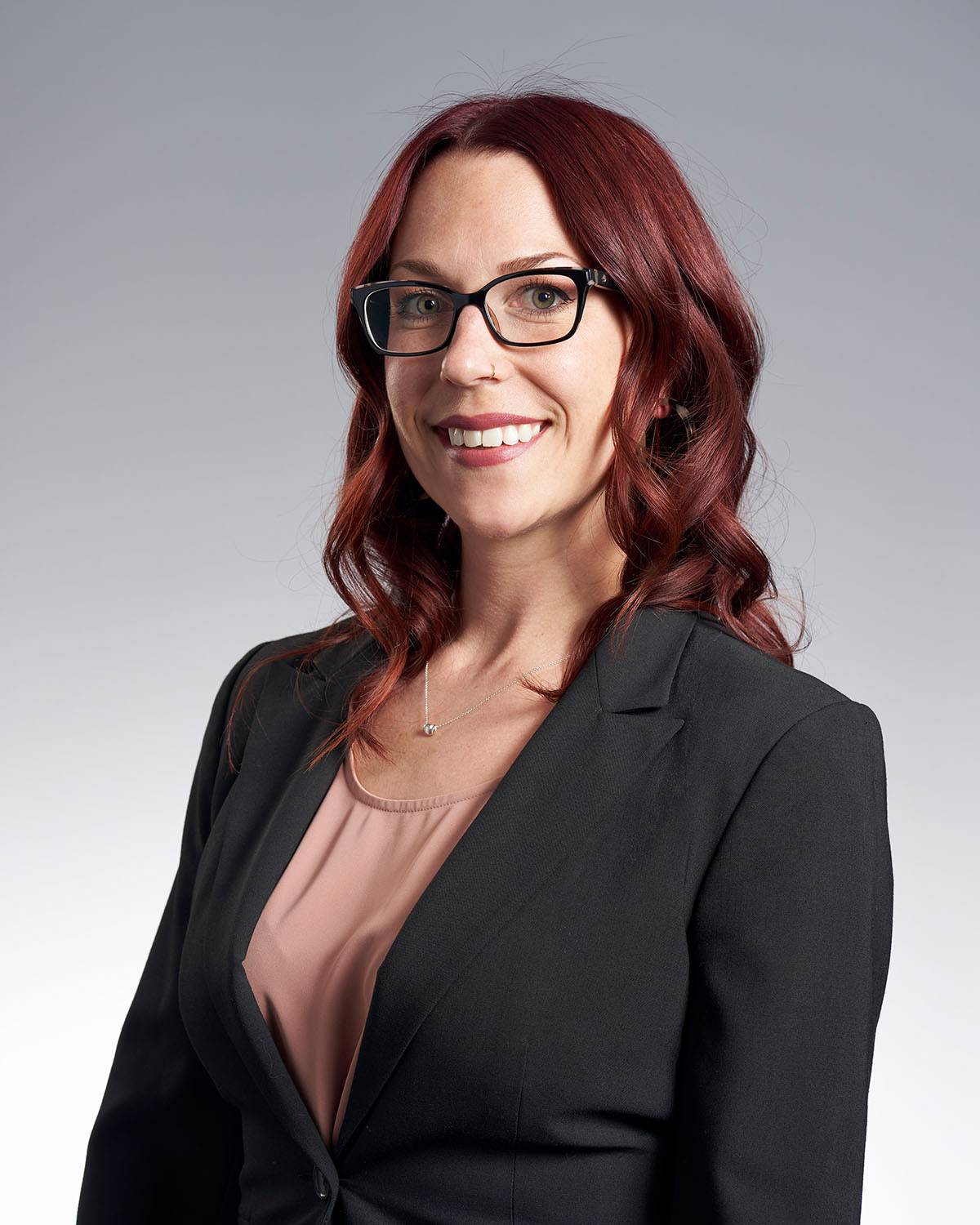 Professional headshot of a woman with red hair and glasses in a dark blazer against a gray gradient background — Headshots by Guffy, San Diego